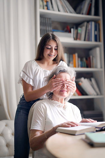 A joyful moment as a young woman playfully surprises her grandmother while reading indoors by covering her eyes. HD phone wallpaper