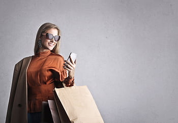 Chic woman in sunglasses browsing on smart with shopping bags indoors. HD wallpaper