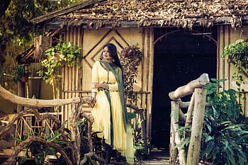 Elegant woman in yellow dress posing by rustic bamboo hut amidst lush greenery. HD wallpaper