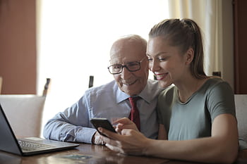 A young woman and an elderly man share a joyful moment using a smart indoors, symbolizing generational connection. HD wallpaper