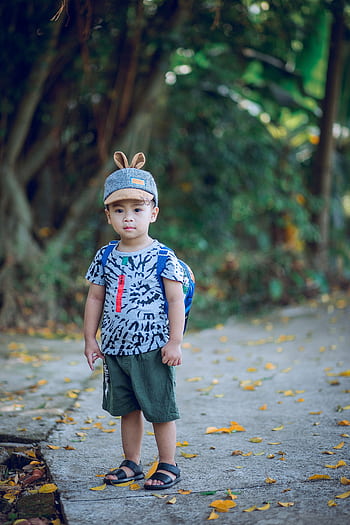 Cute young boy standing outdoors with a backpack, surrounded by trees and fallen leaves. HD phone wallpaper