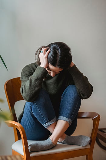 A young woman sitting on a chair indoors, holding her head in distress. HD phone wallpaper