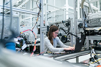 A female engineer focuses on her laptop amidst advanced tech equipment in a lab. HD wallpaper