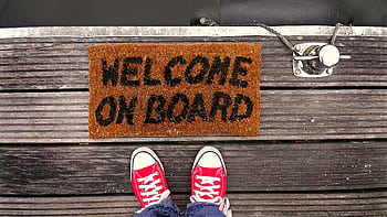 A welcome mat on a wooden dock with a pair of red sneakers visible from above. HD wallpaper