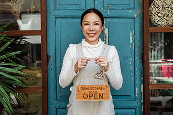 A cheerful woman café owner welcomes customers with a 'we are open' sign at the café entrance. HD wallpaper