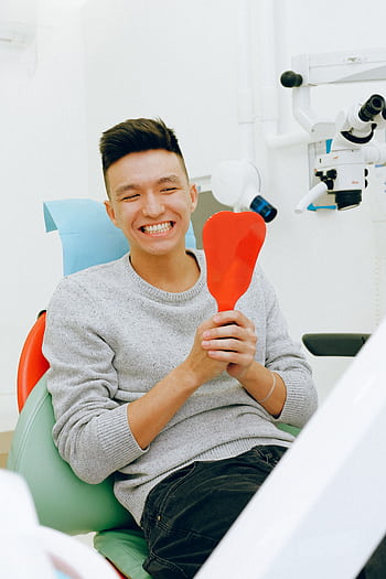 Smiling man holding a mirror during a dental checkup in a clinic setting. HD phone wallpaper