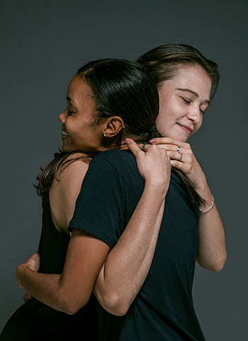 Two women of different ethnicities share a warm hug, symbolizing love and friendship against a gray backdrop. HD phone wallpaper