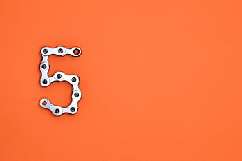 Creative arrangement of a chrome bicycle chain forming the number 5 on a vibrant orange background. HD wallpaper