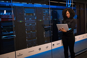 A female engineer using a laptop while monitoring data servers in a modern server room. HD wallpaper