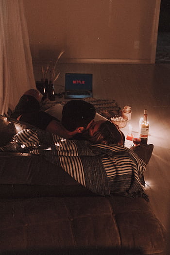 Cozy indoor movie night setup with a couple cuddling on a couch, surrounded by candles and wine. HD phone wallpaper