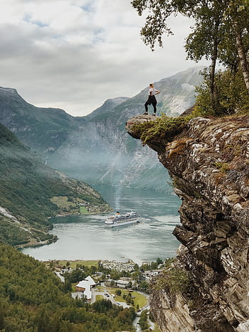 Person enjoys breathtaking view from a cliff above Geiranger Fjord, Norway, with clear skies and cruise ship below. HD phone wallpaper