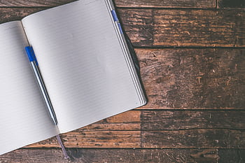 An open notebook with a blue pen lies on a rustic wooden table, inviting creativity and writing. HD wallpaper