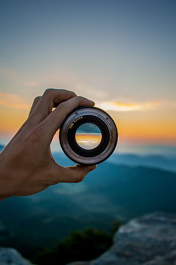 A hand holding a camera lens against a scenic sunset background, capturing nature's beauty. HD phone wallpaper