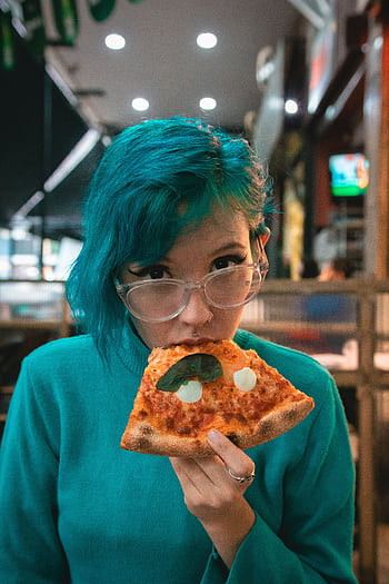 Woman with blue hair and glasses delighting in a tasty pizza slice at an indoor restaurant. HD phone wallpaper