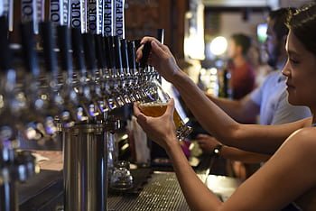 Female bartender pours craft beer in a lively pub setting, creating an inviting atmosphere. HD wallpaper