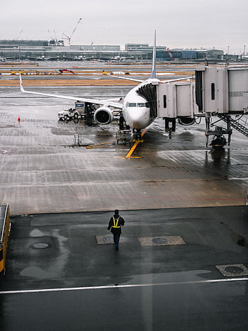 Airplane parked at airport gate on a rainy day with a ground crew member walking. HD phone wallpaper