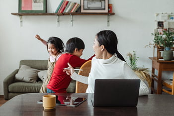 Mother multitasking at home with kids playing in the background, showcasing family dynamics. HD wallpaper