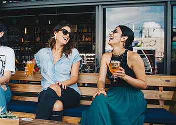 Two women laughing and enjoying drinks outdoors at a trendy bar, creating a vibrant and social atmosphere. HD wallpaper