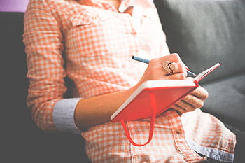 Close-up of a woman writing in a red journal on a sofa, wearing a stylish ring. HD wallpaper