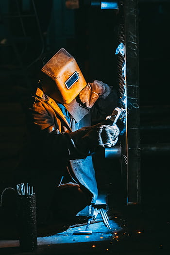 A welder wearing safety gear sparks metal in a dimly lit industrial setting. HD phone wallpaper