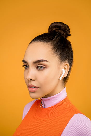 Young woman wearing earbuds, posing against a vibrant orange background. Fashionable and modern. HD phone wallpaper