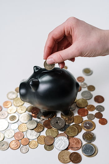 Hand inserting coin into a black piggy bank surrounded by various coins, symbolizing savings. HD phone wallpaper
