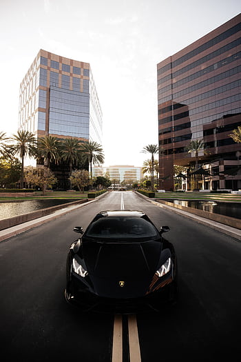 Black Lamborghini driving on a city street flanked by modern skyscrapers. HD phone wallpaper
