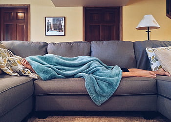 Person napping under a blue blanket on a comfortable couch in a modern living room setting. HD wallpaper