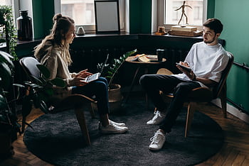 A man and woman engaged in conversation at a stylish, plant-filled home office with modern furniture and digital devices. HD wallpaper