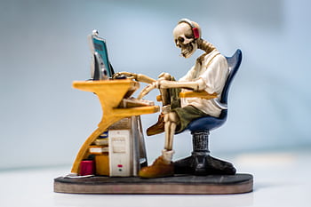 A quirky skeleton figurine wearing heads sits at a desk with a computer, symbolizing burnout. HD wallpaper