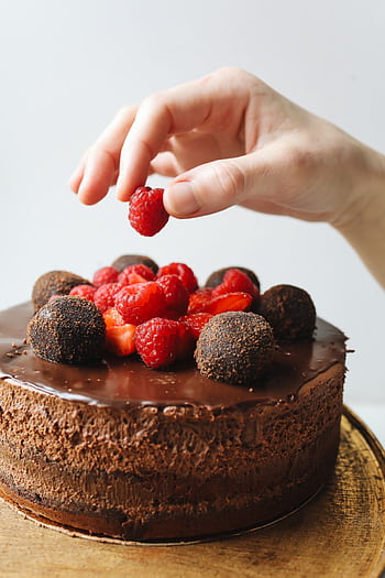 Close-up of a hand decorating a chocolate cake with raspberries and truffles. HD phone wallpaper