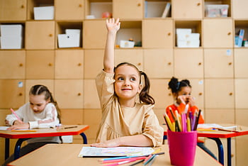Three children engaged in learning activities inside a classroom, with one raising her hand. HD wallpaper