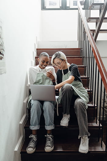 Two diverse women enjoy relaxing and browsing the internet on a laptop while sitting on indoor staircase. HD phone wallpaper