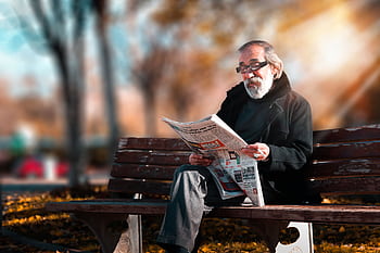 Senior man with beard reading a newspaper on a wooden bench in a sunny park setting. HD wallpaper