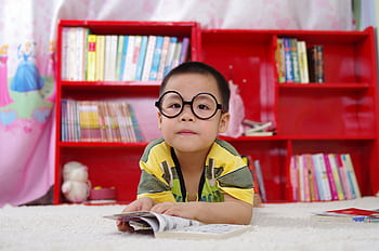 A cute child in glasses reading a book, surrounded by colorful shelves. Perfect shot of childhood learning indoors. HD wallpaper