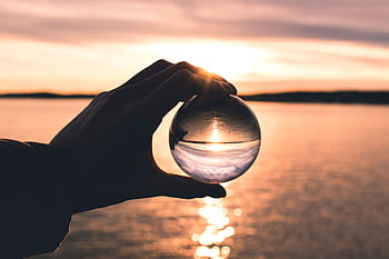 A hand holds a crystal ball reflecting a serene sunset over calm ocean waters. HD wallpaper