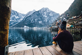 A woman sits on a dock overlooking the serene Hallstatt Lake with snowy mountains in the background. HD wallpaper