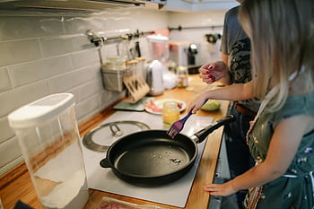 Young girl cooking with a frying pan under adult supervision in a modern kitchen. HD wallpaper