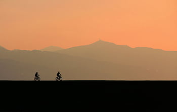 Cyclists on a road at dusk with mountains silhouetted against a vibrant orange sky. HD wallpaper