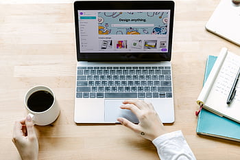 A person using a laptop with design software and holding a coffee at a workspace. HD wallpaper