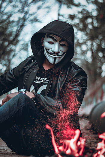 Mysterious figure in mask and jacket sitting by a campfire outdoors. HD phone wallpaper