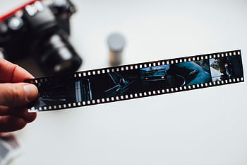 Close-up of a hand holding a vintage film strip with a blurred camera in the background, evoking nostalgia. HD wallpaper