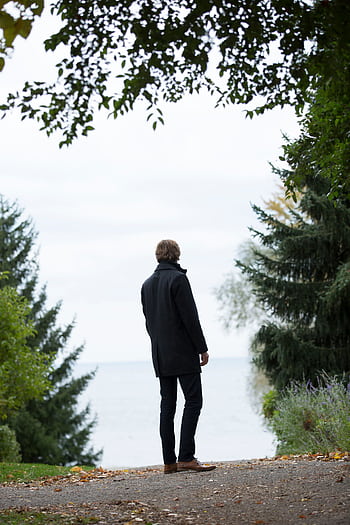 Man in a coat standing on path, surrounded by trees, overlooking a serene lake on a cloudy day. HD phone wallpaper
