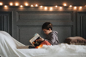 A young boy reads a book in a cozy bedroom setting with warm string lights and soft textures. HD wallpaper