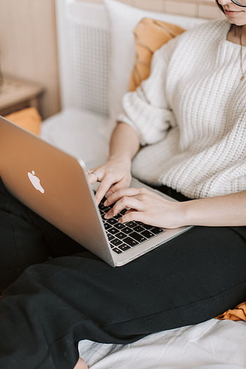 A woman in a sweater typing on a laptop while sitting on a bed in a cozy bedroom environment. HD phone wallpaper