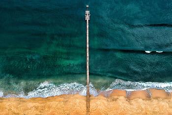 Stunning aerial view of a solitary jetty reaching into the deep blue ocean with vibrant waves crashing ashore. HD wallpaper