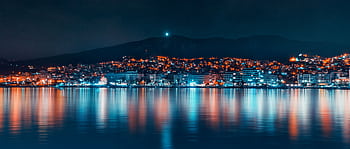 A tranquil cityscape at night with vibrant lights reflecting on the calm waterfront. HD wallpaper