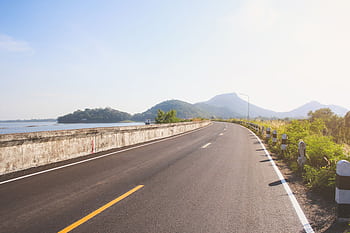 A que empty road curving alongside a tranquil lake, framed by lush mountains under a clear sky. HD wallpaper