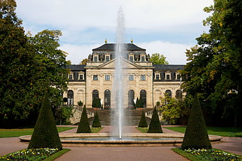 Elegant historic mansion facade with central fountain surrounded by lush gardens, offering a glimpse of classic architecture. HD wallpaper