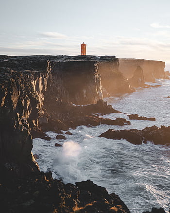 Stunning view of a lighthouse on Iceland's rugged coastline during sunset, showcasing dramatic cliffs and ocean waves. HD phone wallpaper
